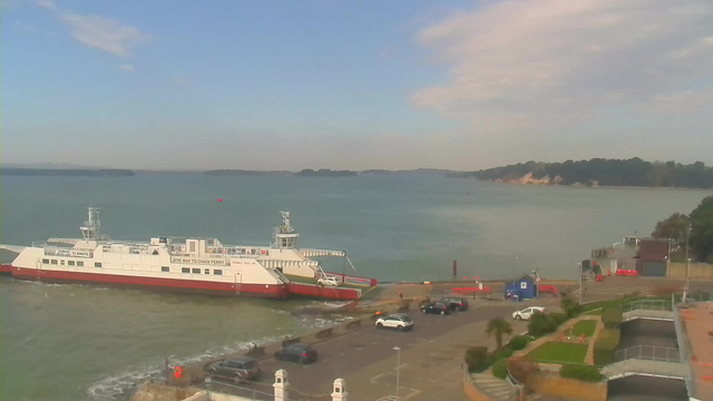 A ferry is docked at a pier with a view of calm water extending towards the horizon. The background features a distant shoreline with trees and cliffs. In the foreground, there is a partially paved area with several parked cars. A small blue structure and a few red objects are visible nearby. The sky is mostly clear with scattered clouds.