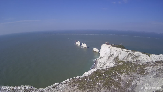 A panoramic view of a coastal landscape featuring white chalk cliffs. In the distance, two lighthouses are situated on rock formations jutting out into the blue sea. The sky above is clear with minimal clouds, and the ocean displays gentle waves. The foreground includes rocky terrain with sparse vegetation.