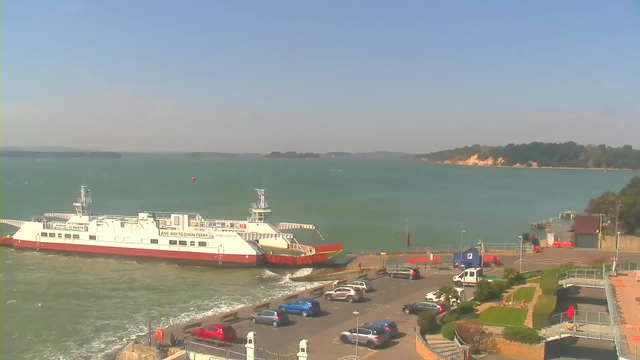 A ferry is docked at a pier beside a body of water, with waves gently lapping against the structure. Various parked cars are visible in a nearby parking lot, and there are green trees and a sandy beach in the background. The sky is clear and blue, indicating a sunny day. There are some structures and pathways along the shoreline, and the distant land features hills and trees.