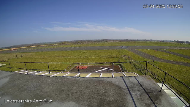 A clear blue sky stretches above a grassy airfield, where a light aircraft is seen taxiing along a runway. In the foreground, a flat rooftop railing frames the image, and a concrete area with a white cross outline can be seen to the left. The landscape is dotted with a few wooden fencing sections, and a few distant hills are visible in the background. The image captures a peaceful, sunny day at a small airport.