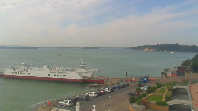 A ferry with a white upper body and a red lower hull is docked by a waterfront, with gentle waves in the foreground. In the background, there are hills and a blue sky with soft clouds. Several cars are parked along the waterfront, and a small building is visible on the right side. The overall scene conveys a calm, sunny day by the water.