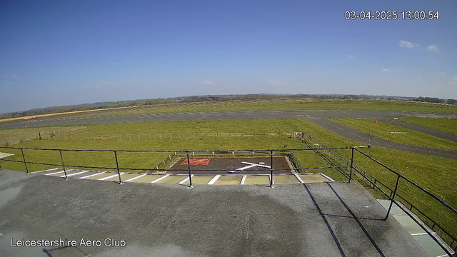 A wide view from a rooftop at Leicestershire Aero Club shows an expansive green field with a clear blue sky. A portion of the grass is marked with a red symbol and a white "X." In the distance, a runway is visible with light-colored pavement contrasting against the green grass. A fence lines the area, and the scene is bright and sunny, indicating a lovely day.