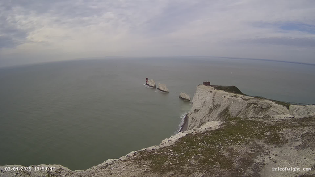 A panoramic view of a coastline featuring steep white chalk cliffs and a grayish-green sea. In the distance, there are two rock formations protruding from the water, with a red and white striped lighthouse on one of the rocks. The sky is partly cloudy with a mix of light blue and gray. In the foreground, the rocky coast is visible, scattered with vegetation.