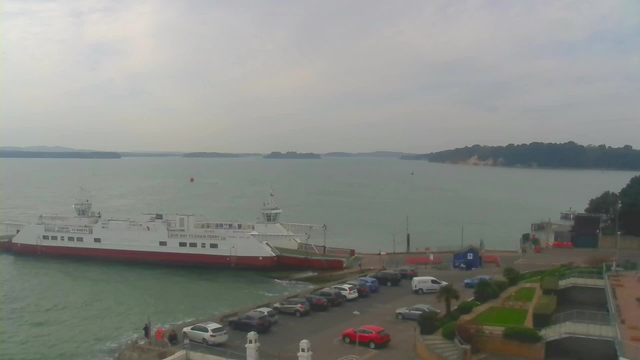 A white ferry with a red bottom is docked at a pier, surrounded by water. Several cars are parked in a lot nearby, with a mix of colors including silver, blue, and red. In the background, a rocky shoreline with trees can be seen, along with hills in the distance. The sky is overcast, creating a muted light over the scene.