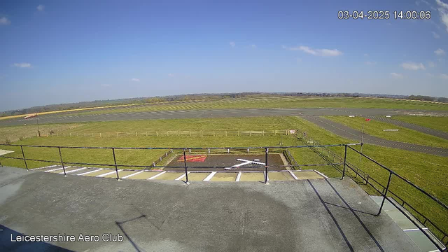 A clear blue sky with a few scattered clouds forms the backdrop of this landscape view from a webcam. In the foreground, a balcony railing is visible, while below is a grassy area with a helicopter landing pad marked by a red square and a white cross. Beyond the pad, there is an expansive green field leading to a runway, which is mostly clear and bordered by a wooden fence. The horizon shows a gentle rise in the terrain, indicating distant hills.