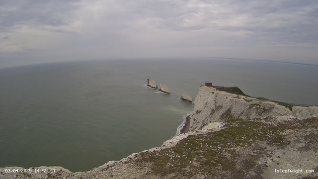 A view of a coastline featuring white chalk cliffs overlooking a calm sea. In the distance, there are several rocky islets, with a red-and-white lighthouse on the furthest islet. The sky is cloudy with varying shades of gray, and a grassy area is visible in the foreground of the cliffs. The sea appears calm and is a muted blue-green color.