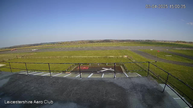 A clear blue sky is seen above a grassy area with an airstrip. In the foreground, there is a structure with a railing, indicating an elevated viewpoint. Below, the runway is visible, along with a small airplane positioned to the side. Fencing borders the grassy fields, and there is no visible activity on the runway. The overall scene reflects a sunny day at an airfield.