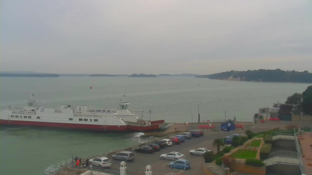 A grey, overcast sky is visible above a calm body of water, where a large white ferry with a red lower hull is docked near a shoreline. There are several parked cars in the foreground, with a few people on the dock near the ferry. In the background, multiple small islands can be seen on the horizon, and some greenery lines the shoreline. The scene appears tranquil, with no visible movement on the water's surface.