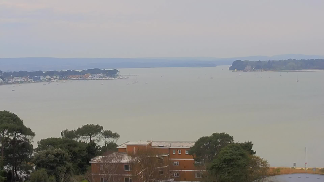 A serene view of a calm body of water stretches across the image, with distant hills visible under a pale sky. In the foreground, there are trees and a brick building, while several small boats dot the water's surface. The shoreline is lined with white buildings and docks, indicating a coastal area, and the scene conveys a peaceful atmosphere.