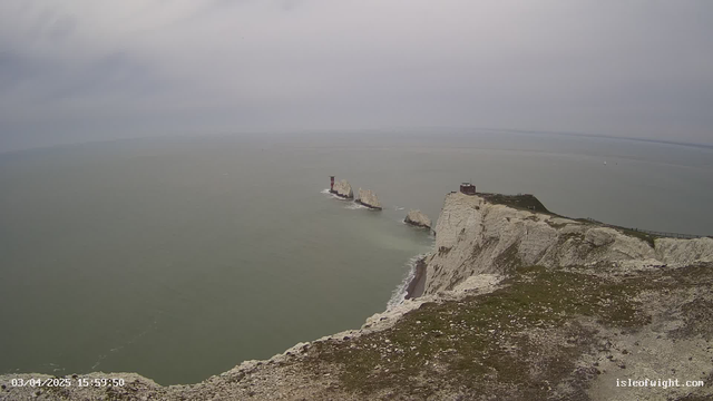A coastal scene featuring steep white cliffs rising above a calm sea. In the foreground, patches of green grass are visible. In the distance, several rocky formations emerge from the water, with a brick lighthouse standing prominently on one. The sky is overcast, and the atmosphere appears gray and hazy. The image captures a tranquil yet rugged natural landscape.