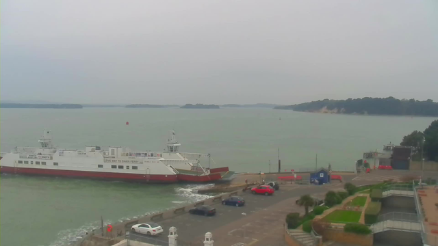 A large white and red ferry is docked at a pier, with water gently lapping against the side. In the background, there are several small islands visible on the horizon under a gray sky. The foreground features a parking area with a few cars, including a white one and a red one. There are green shrubs and landscaped areas along the waterfront, with pathways leading toward the pier.