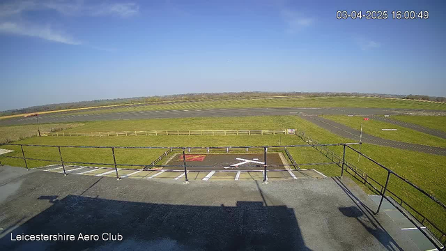 A clear sky with a few clouds is visible overhead. The scene shows a grassy area surrounding a runway at the Leicestershire Aero Club. In the foreground, a helipad is marked with a large white X on a dark surface, and there are white lines indicating the boundaries. A wooden fence outlines the grassy area, and part of a taxiway can be seen in the distance. The setting is bright and open, typical of an airfield environment.