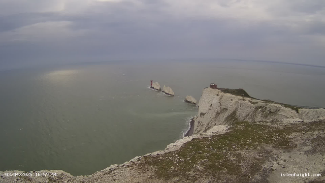 A coastal view depicting white chalk cliffs that rise sharply from the sea. The ocean is a muted green color with gentle waves. On the left, there is a series of rocky formations emerging from the water, the furthest one featuring a red and white striped lighthouse. Above, the sky is overcast with gray clouds, creating a dramatic atmosphere. A grassy area is visible in the foreground, adding a touch of greenery to the scene.