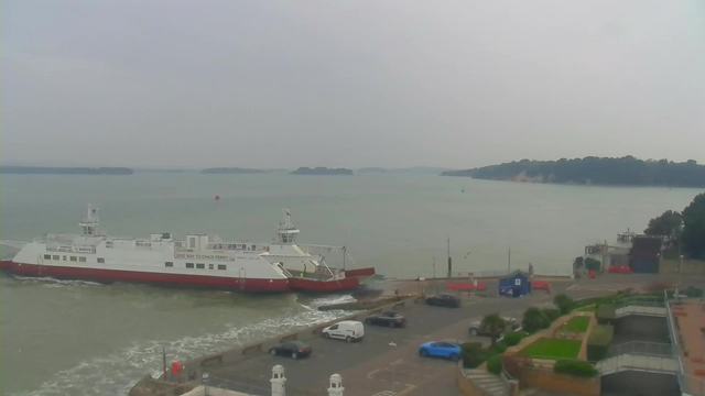 A ferry with a white body and red bottom is approaching a terminal by the water, where it is preparing to dock. The background features a calm body of water with several small islands visible in the distance under a cloudy sky. The foreground includes a road with parked vehicles, including a white van and a blue car, along with a landscaped area with greenery and a few people walking around the terminal.