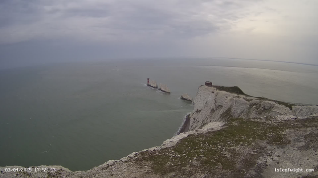 A coastal view featuring white chalk cliffs that rise sharply along the edge of the ocean. In the distance, a series of rocky outcrops are visible, with a red and white striped lighthouse positioned prominently on one. The sky is overcast, with grey clouds covering most of the horizon, while the sea appears calm and stretches out to meet the sky. A building is situated near the cliff's edge on the right side of the image.