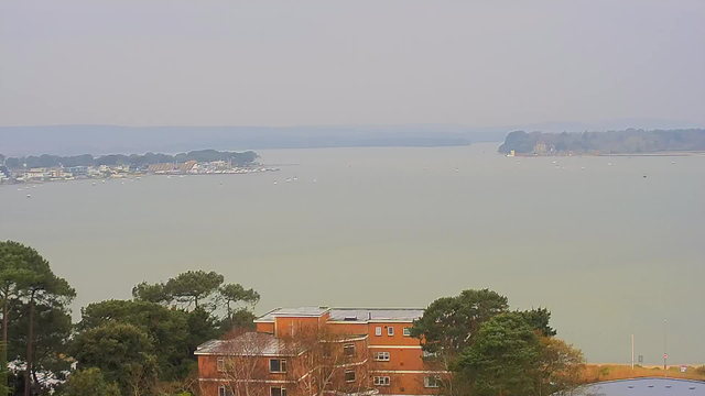 A panoramic view of a calm body of water, with small boats scattered across it. The shoreline features a cluster of buildings, primarily brick-colored, along with trees in the foreground. In the background, the water extends towards distant hills and a shoreline that includes more buildings and greenery. The sky is overcast, creating a subdued light across the scene.