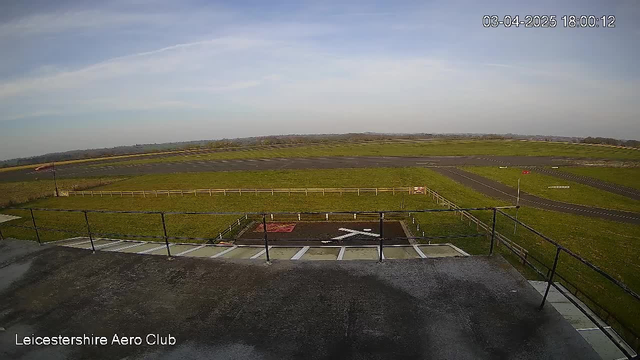 A view from a webcam at the Leicestershire Aero Club, showing a wide open space with a grassy area and a runway. The sky is mostly clear with some clouds. In the foreground, there is a railing and a helipad marked with a red cross. To the right, there is a runway with a few markings and a small red sign. The scene captures the calm atmosphere of an airport environment.