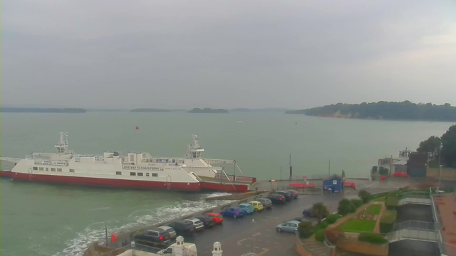 A ferry is docked at a jetty, surrounded by water that appears calm and slightly greenish. The ferry is mostly white with a red bottom and has text on its side, reading "GIVE WAY TO CHAIN FERRY." Several parked cars in various colors—blue, red, yellow, and gray—line the road adjacent to the parking area. There are some trees and hills in the distance, under a cloudy sky. The atmosphere looks overcast, suggesting it might be a gloomy day.