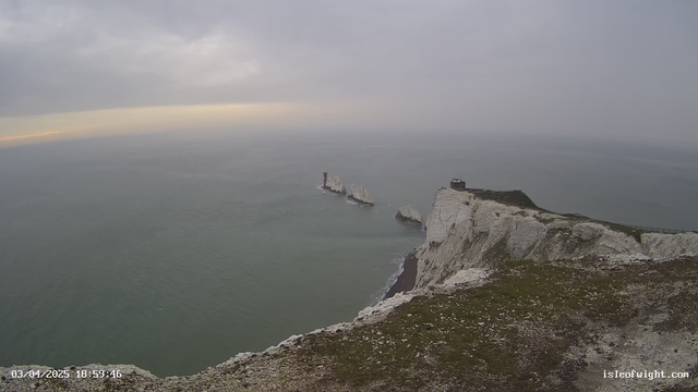 A scenic view of the coastline featuring white chalk cliffs that rise sharply above the sea. In the distance, a lighthouse stands on a rock formation surrounded by water. The sky is overcast with gray clouds, and the light appears dim, indicating a possibly cloudy or sunset setting. The foreground shows grassy terrain with small rocks scattered about.