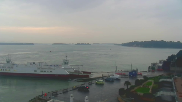 A cloudy sky looms over a harbor scene with a ferry docked at a pier. The ferry has a white body with a red bottom and is labeled "CHAIN FERRY." Several vehicles, including a yellow car and a white van, are parked near the water's edge. The water is calm with visible ripples, and a few distant islands can be seen on the horizon. A small building with a tent is situated to the right of the pier, surrounded by greenery.