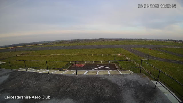 A view of an airfield from a high vantage point. The foreground features a flat rooftop surface with a railing. In the middle ground, a grassy runway stretches across the scene, bordered by a white wooden fence. To the right, there are two paved taxiways, and a red sign is visible near the edge of the runway. The sky is mostly clear with soft, light-colored clouds. The timestamp in the top right corner indicates the image was taken on April 3, 2025, at 19:00:46.