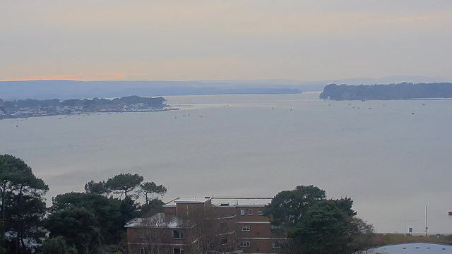 A wide view of a calm body of water extending towards the horizon, with a light gray sky overhead. In the foreground, there are some trees and a building, while various small boats are scattered across the water. On the left side, there is a shoreline with several structures and houses visible. The distant hills create a gentle backdrop against the water.