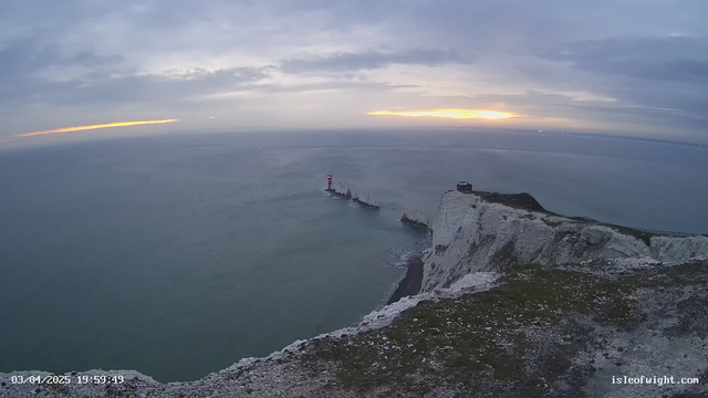 A coastal view at sunset, showing a calm sea with gentle waves. In the foreground, there are steep, white chalk cliffs with some green grass and rocks. To the right, a small lighthouse stands on a outcropping of rocks, illuminated with a red light. The sky is partly cloudy, with hints of orange and yellow near the horizon as the sun sets. The scene conveys a tranquil atmosphere.