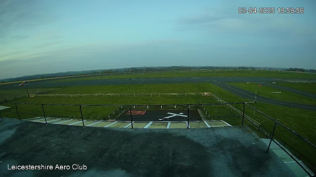 A clear sky is visible above a large, open grass field designed for aviation activities. In the foreground, there is a helipad marked with a white "X" on a black surface. Surrounding the pad is a white fence. A small wind sock can be seen on the left, indicating wind direction. In the distance, the field transitions to asphalt runways and other airfield infrastructure. The image is taken from an elevated perspective, looking out over the aerodrome.