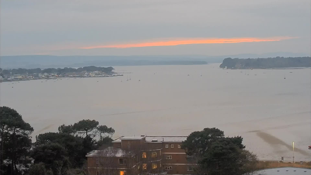 A coastal landscape during twilight. The sky is overcast with soft gray clouds, and a hint of orange is visible along the horizon, suggesting sunset. In the foreground, there are trees partially obscuring a brown building with illuminated windows. The water stretches out into the distance, dotted with small boats. On the opposite shore, various buildings and trees line the waterfront, with hills visible in the far background.
