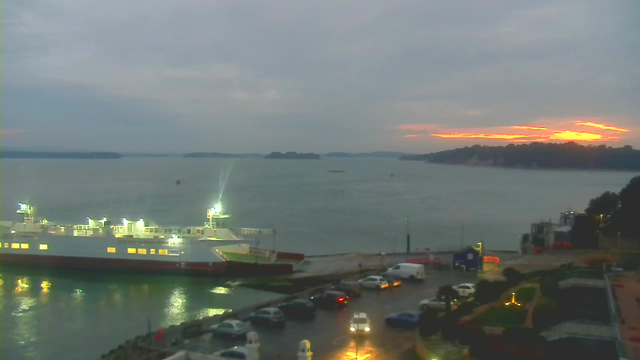 A calm waterfront scene just before sunset. In the foreground, a large ferry is docked at a pier, lit by several bright lights. A parking lot is visible with a lineup of cars, some with headlights on. The water is gentle and reflects the subdued light, while the sky above is mostly cloudy, with hints of orange and pink near the horizon, indicating the setting sun. On the right, there’s a small harbor area with additional boats, and further in the background, distant hills are partially obscured by clouds.