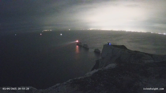 A nighttime coastal scene with a prominent cliff edge on the right side. The ocean is visible with some light reflections, while several boats are gently illuminated in the dark water. A lighthouse beam shines out to the left, showcasing a red light. The sky is overcast, and faint city lights can be seen in the distance. The date and time are displayed at the bottom left corner of the image.