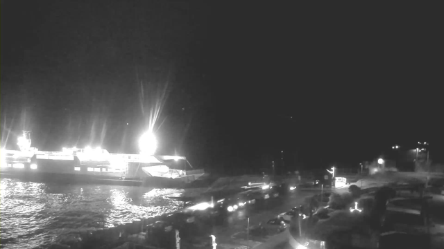 A nighttime scene showing a large ferry docked at a pier with bright lights illuminating the area. The water is dark, reflecting the light from the ferry and nearby streetlights. Various vehicles, possibly cars, are parked along the waterfront. The overall atmosphere appears calm and quiet.