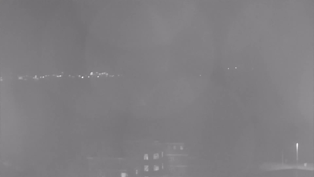 A foggy or low-light scene showing distant lights faintly visible on the horizon. Foreground includes outlines of buildings with some illuminated windows. The overall atmosphere appears hazy, creating a blurred effect.