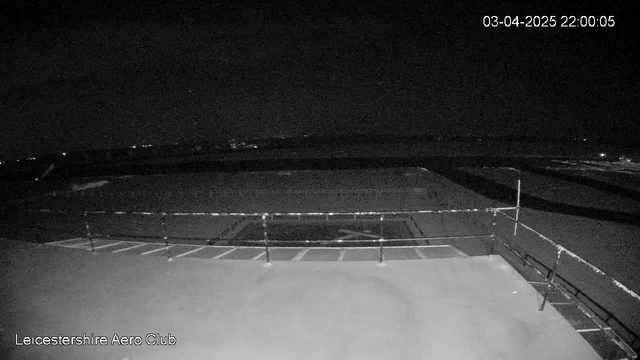 A dark scene captured from a webcam at night, showing a flat rooftop with a railing along the edge. There are faint lights visible in the distance, indicating a possibly urban environment. The date and time are displayed in the top right corner. Overall, the setting appears quiet and still with minimal visible activity.
