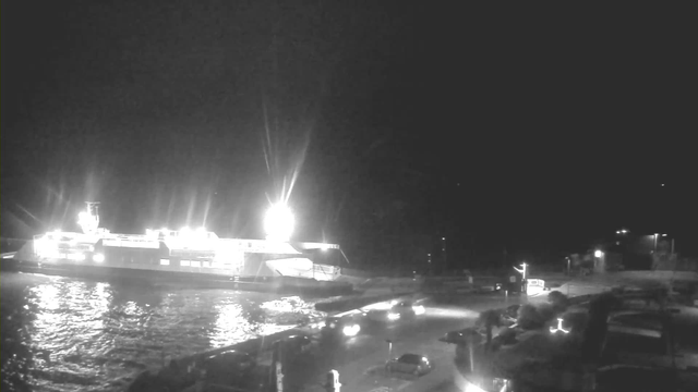 A nighttime scene showing a ferry docked near a waterfront. The ferry is brightly lit and reflects on the dark water. Nearby, several cars are visible on the road, with some streetlights illuminating the area. The background is mostly dark with limited visibility of surrounding structures.