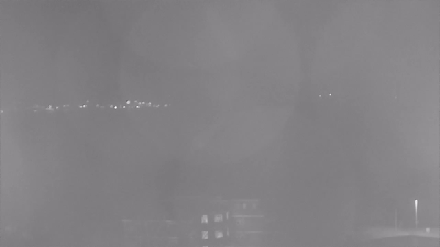 A dimly lit scene with a grayish, foggy background. In the distance, there are faint, blurred lights that seem to be scattered across a horizon, possibly representing a city or buildings. The foreground appears dark, with a few indistinct shapes at the bottom, suggesting a structure or surface. The overall atmosphere is unclear and obscured, giving a sense of low visibility and dampness.