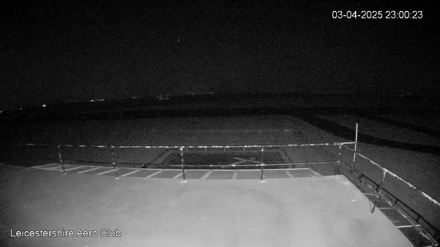 A dark, nighttime scene captured by a webcam, showing a flat surface with a railing at the bottom of the image. In the distance, there are faint lights, possibly from a nearby town or aircraft, and an open area that suggests a runway or large field. The date and time are displayed in the upper right corner, indicating it is 11:00 PM on April 3, 2025. The overall visibility is low due to the darkness.
