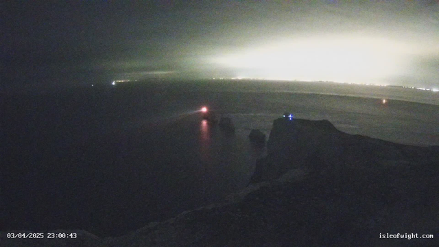 A dimly lit coastal scene at night, featuring rocky cliffs extending into the ocean. On the left side, a red light from a boat is visible, gliding on the water's surface. To the right, a blue light can be seen, possibly from a structure on the cliffs. The background shows a faint glow from lights on the horizon, suggesting a distant town or city. The overall atmosphere is quiet and serene, with dark tones dominating the image.