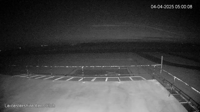 A dark, nighttime scene captured by a webcam showing a flat surface with a railing on the right side. In the background, there are faint lights that suggest distant buildings or vehicles. The image is predominantly dark, with minimal light visible. The date and time in the top corner indicate April 4, 2025, at 5:00:08 AM.