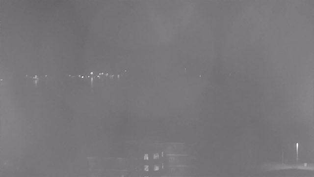 A dimly lit scene showing a blurry view of a body of water with faint reflections of lights along the surface. In the foreground, a building is partially visible, with some windows illuminated. The overall image has a grayish tint, suggesting low visibility conditions possibly due to fog or haze.