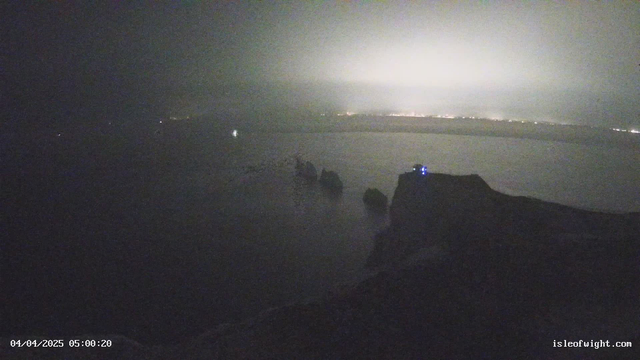 A dimly lit coastal scene captures a foggy early morning at a cliffside location. In the foreground, a dark rock formation juts into a calm sea. A small building with blue lights is situated near the edge of the cliff. The background reveals a hazy horizon with faint lights from a distant area, suggesting a city or town. The atmosphere is quiet and serene, with a muted color palette dominated by grays and blues.