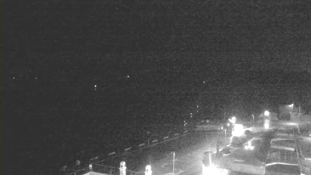 A dimly lit scene captured from a webcam shows a waterfront area at night. In the foreground, there are faint outlines of structures, possibly buildings or piers, with some illuminated spots likely representing lights or vehicles. The background appears mostly dark, with a few distant lights visible on the horizon, suggesting the presence of water or an open space beyond the immediate area.