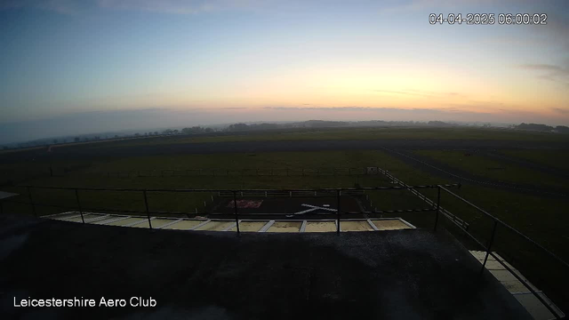 A view from a webcam at the Leicestershire Aero Club showing a wide, open field at dawn. The sky has soft pastel colors of light blue and pink, indicating sunrise. In the foreground, there is a railing and part of a structure, while the horizon includes distant trees and a misty landscape. The ground is mostly flat with some dark patches indicating runway areas. The scene conveys a tranquil morning atmosphere.