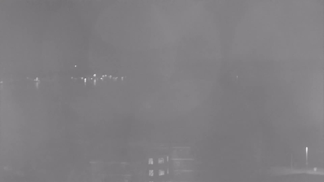 A foggy night scene captured from a webcam, showing indistinct outlines of a body of water in the background. Faint lights reflecting on the water are visible, along with blurry shapes of buildings or structures in the foreground. The overall image has a grayish tone, making details difficult to discern.
