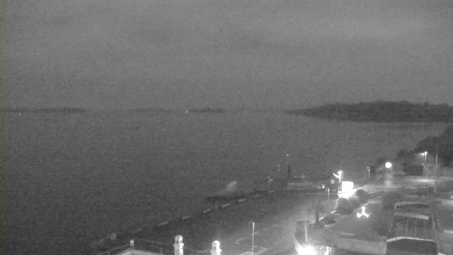 A dark, cloudy scene featuring a calm body of water extending towards the horizon. In the foreground, there is a dock or pier with a few blurred structures and faint lights. To the right, there are boats parked and some illuminated areas that suggest buildings or facilities nearby, while distant land forms an outline along the horizon. The overall atmosphere is quiet and somewhat gloomy due to the low light conditions.