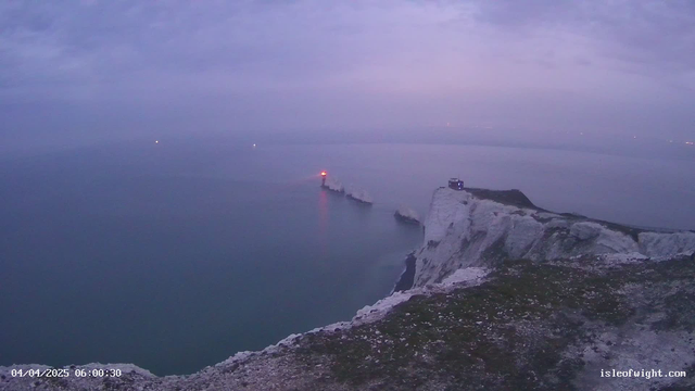 A misty seascape captures a rocky coastline with chalk cliffs. A lighthouse with a red light stands at the tip of a jutting outcrop. The sky is overcast and pale blue, while the calm sea reflects this soft light. In the distance, faint lights can be seen on the horizon, hinting at boats or distant land. The foreground shows grassy terrain leading to the cliffs. The date and time in the bottom corner indicate it is early morning.
