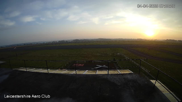 A panoramic view of a grassy field under a cloudy sky at dawn. The sun is rising on the horizon, creating a warm orange glow. In the foreground, there is a railing, with a portion of a white structure visible on the ground below. The field stretches out with a runway marked in dark asphalt and some distant trees. The timestamp in the top right corner shows the date and time.
