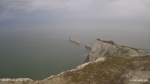 A coastal scene featuring white chalk cliffs on the right, with a flat landscape of grass and scattered rocks. To the left, there are several rocky formations jutting out into a calm sea. In the distance, there is a lighthouse with red and white stripes on one of the rocks, and a small building on top of the cliffs. The sky is overcast with grey clouds, creating a muted and serene atmosphere.