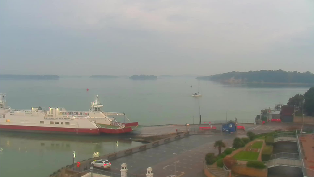 A serene waterfront scene features a large white and red ferry docked at a pier surrounded by calm water. The background includes several small islands and boats on the water. A partially cloudy sky reflects a soft light over the landscape. In the foreground, there is a parking area with a few cars and some greenery, including patches of grass and shrubs. The overall atmosphere is tranquil and slightly overcast.