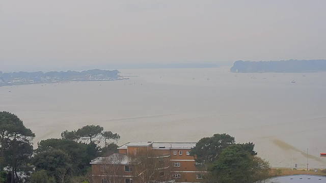 A view of a calm body of water under a cloudy sky. In the foreground, there are several trees and a few buildings with orange brick exteriors. In the distance, a shoreline lined with trees can be seen, along with a few boats scattered across the water. The horizon appears hazy, indicating a distant landmass. The overall scene conveys a serene, overcast day by the water.